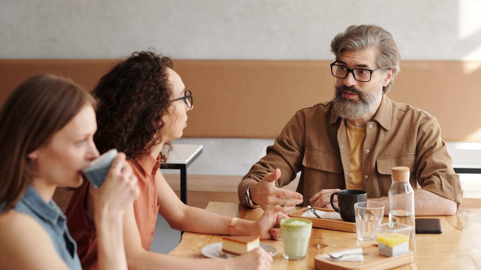 Três adultos de idades diferentes em conversa à volta de uma mesa, um deles a meio de uma frase