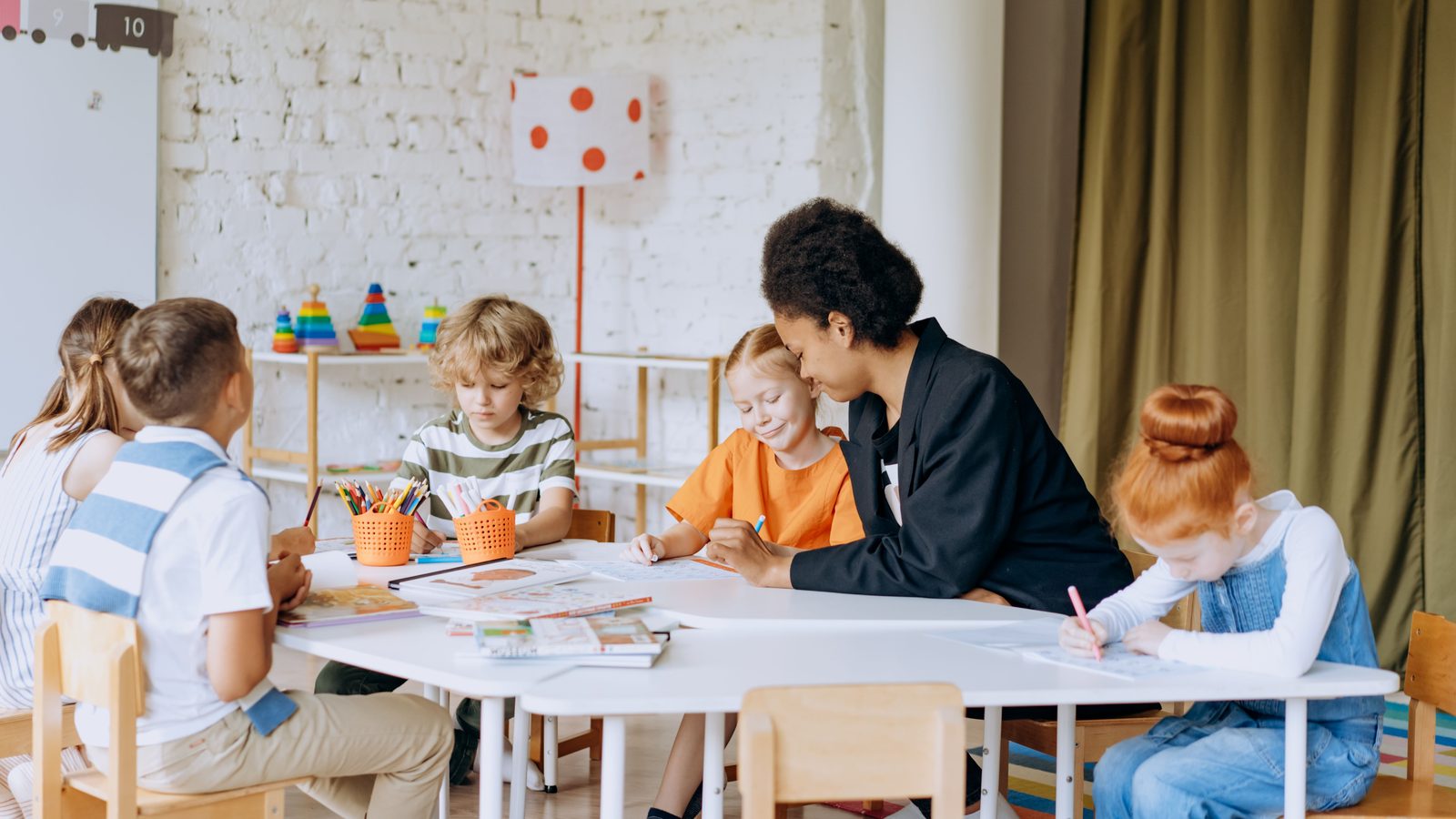 Cinco crianças e uma professora à volta de uma mesa branca, a desenhar e a trabalhar num projeto, com potes de lápis de cor
